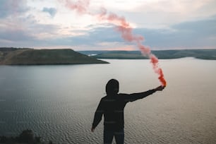 Stilvoller Hipster mit roter Rauchbombe in der Hand, auf dem Felsenberg mit herrlichem Blick auf den Sonnenuntergang am Fluss. Atmosphärischer Moment. Reisender Typ mit Rauch. Speicherplatz kopieren