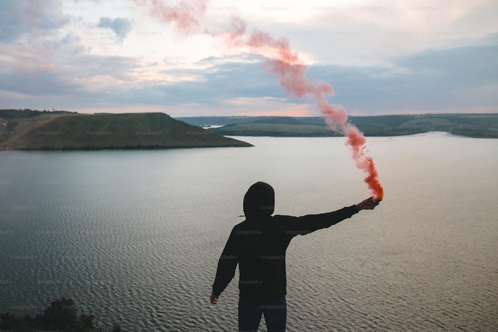 赤い発煙筒を手にしたスタイリッシュなヒップスターが、川の素晴らしい夕日の景色を望む岩山の頂上に立っています。雰囲気のある瞬間。煙を吐く旅人の男。コピースペース