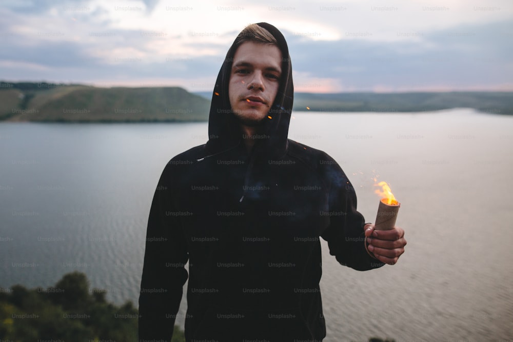 Ultras hooligan holding flare torch with burning fire in hand while standing on top of rock mountain with amazing sunset view on river . Atmospheric moment. Copy space