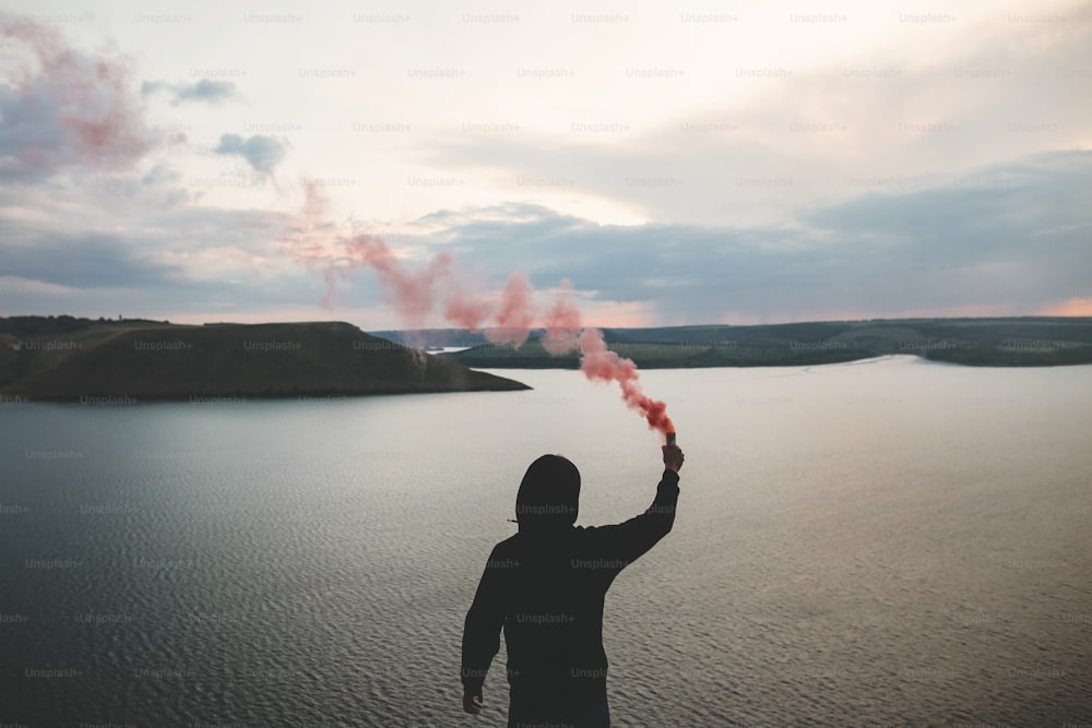 Stylish hipster holding red smoke bomb in hand, standing on top of rock mountain with amazing sunset view on river. Atmospheric moment. Traveler guy with smoke. Copy space