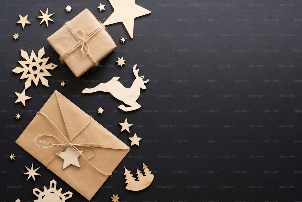Adornos navideños de madera, caja de regalo, sobre de papel kraft con carta sobre fondo negro oscuro. Maqueta de tarjeta de felicitación navideña. Composición mínima de estilo plano, vista superior, espacio de copia.