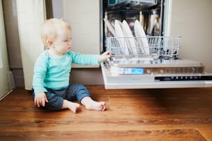 Little child helping to unload dishwasher. Baby girl sitting on the floor in the kitchen. Little child at home