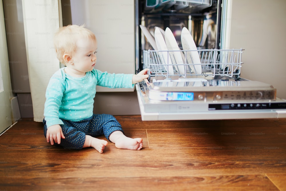 Kleines Kind hilft beim Entladen der Spülmaschine. Kleines Mädchen sitzt auf dem Boden in der Küche. Kleines Kind zu Hause