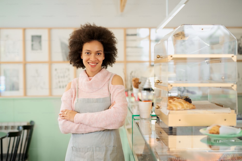 Beautiful smiling mixed race female owner of pastry shop dressed in apron standing with arms crossed in shop.