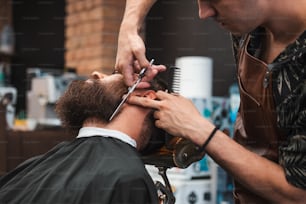 理髪店の肘掛け椅子に座り、美容師がハサミで髭をつまずかせる髭を生やした男性。理髪店の男にあごひげを刈る床屋の接写