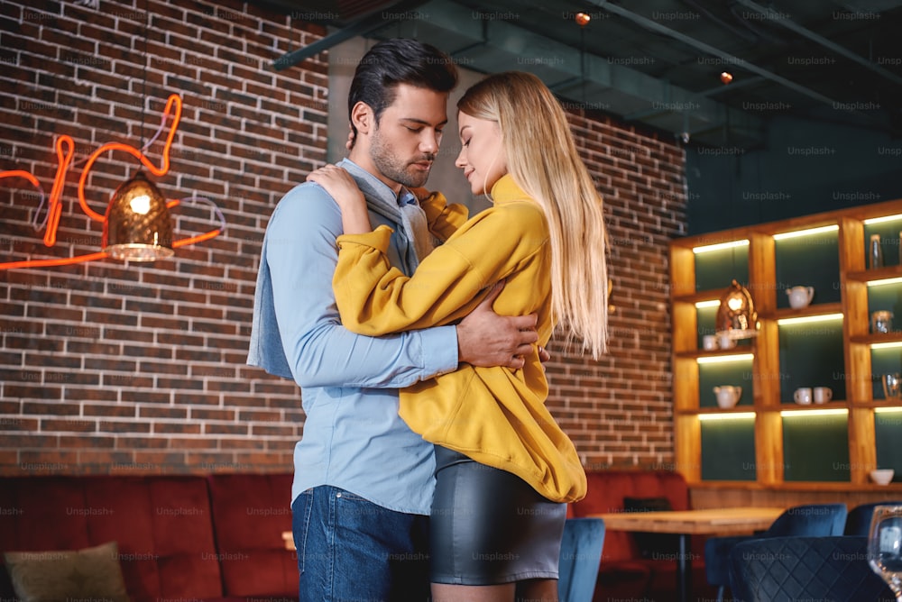 カジュアルな服装の男女がレストランで立って抱き合っている肖像画。ロマンチックなディナーのコンセプト。水平ショット
