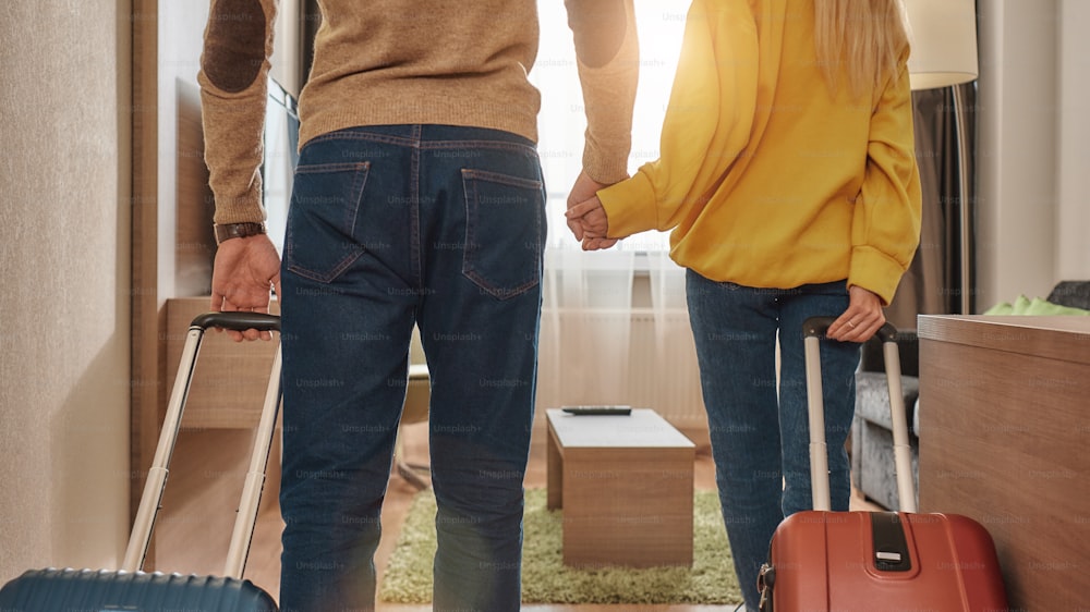 Cropped portrait of young man and woman in casual wear with suitcases entering their room in a hotel. They are holding each other's hands. Travelling together concept. Horizontal shot. Rear view