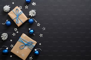 Christmas gifts with blue ribbon, blue balls, fir tree branches, cones, confetti on dark black background with copy space. Flat lay, top view, overhead. Christmas holidays, New Year banner mockup.