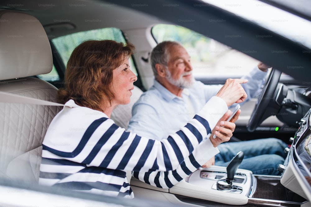 A happy senior couple with smartphone sitting in car, talking.