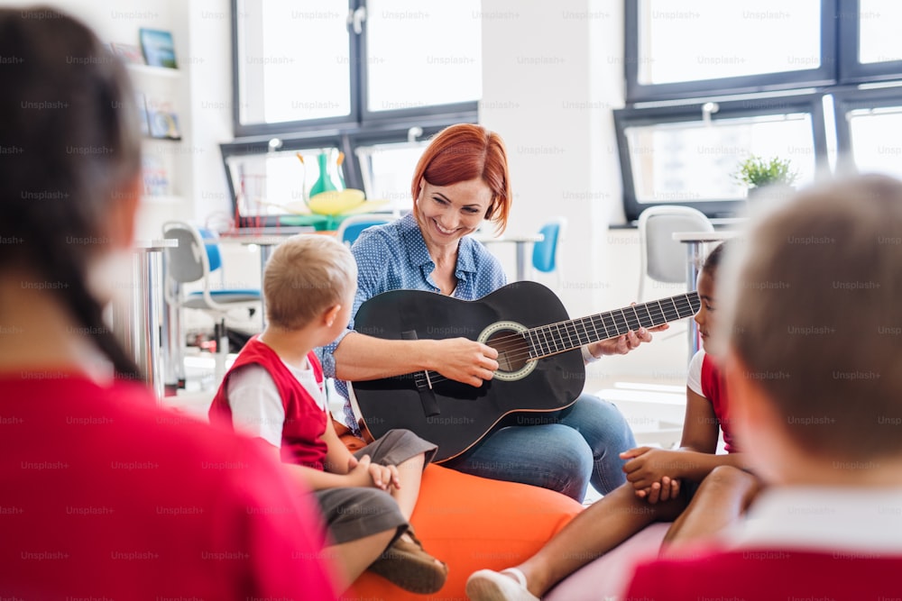 A group of small school kids and teacher with guitar sitting on the floor in class, singing.