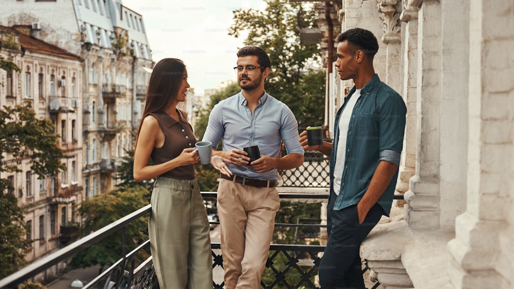 Conversation. Young cheerful colleagues in casual wear holding cups and talking with each other while standing on the balcony. Coworkers resting. Coffee break