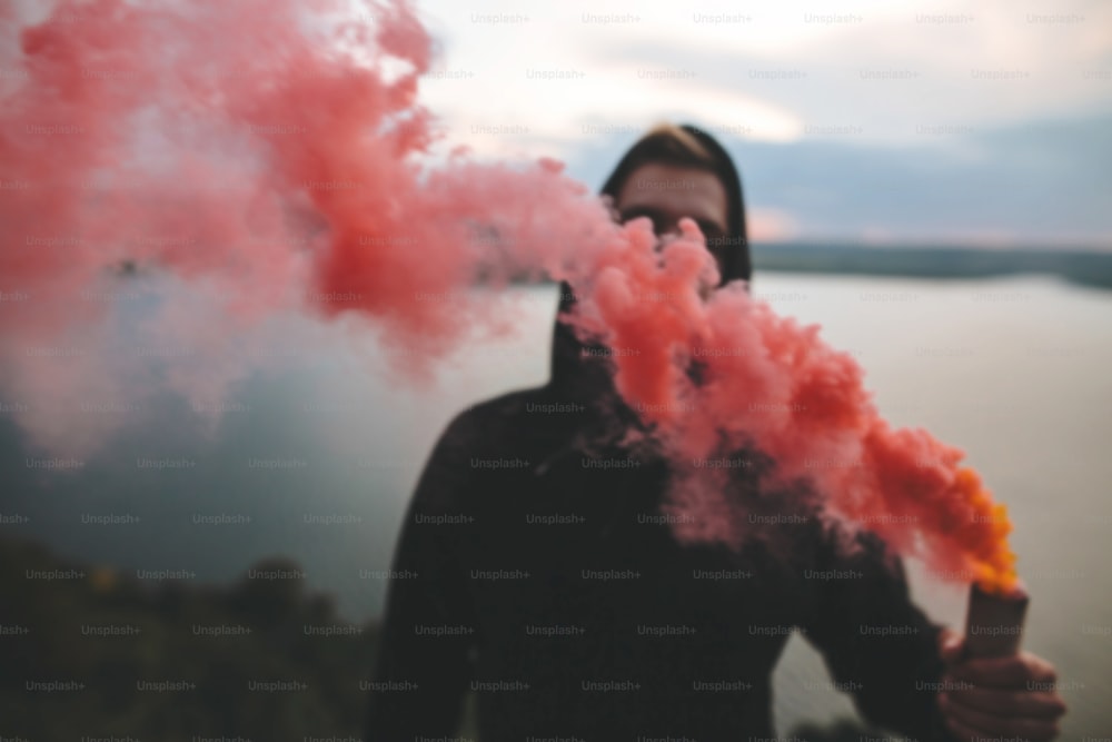 Immagine sfocata di un teppista ultras che tiene in mano una bomba fumogena rossa, in piedi sulla cima di una montagna rocciosa con una vista mozzafiato sul fiume. Momento atmosferico. Spazio di copia