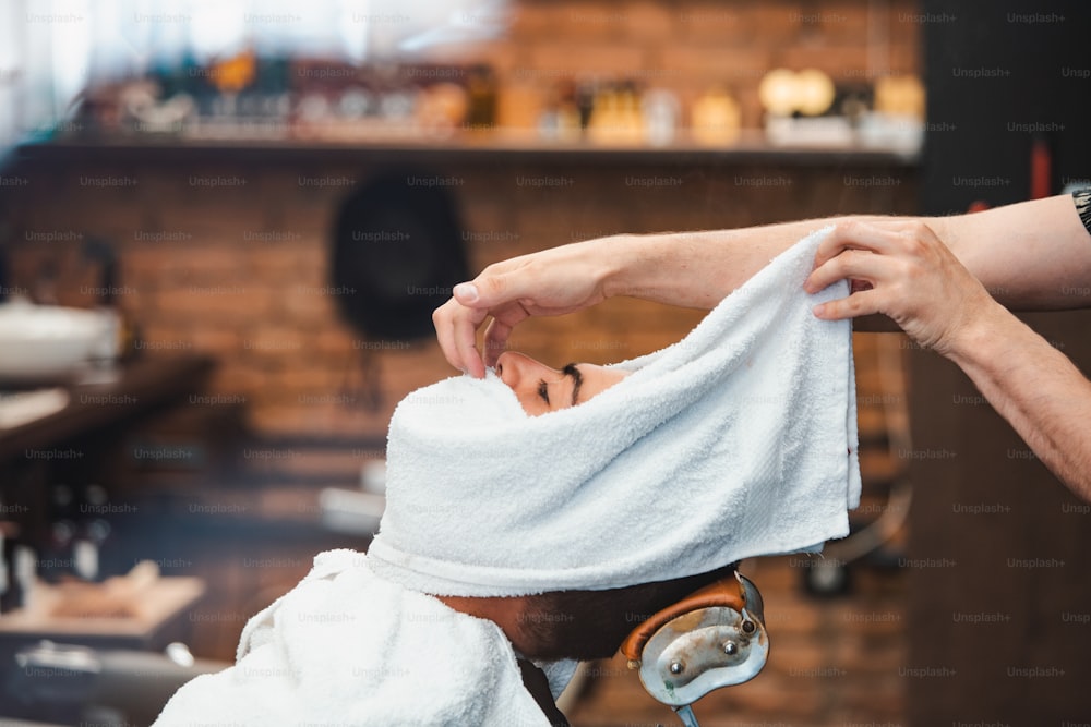 Barbier bedeckt das Gesicht eines Mannes mit einem heißen Handtuch. Traditionelles Ritual, den Bart mit heißen und kalten Kompressen in einem alten Friseursalon zu rasieren. Kundin mit heißem Handtuch im Gesicht vor der Rasur im Friseursalon