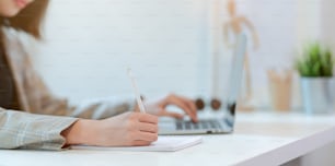 Close-up view of young businesswoman typing on laptop computer and writing on notebook