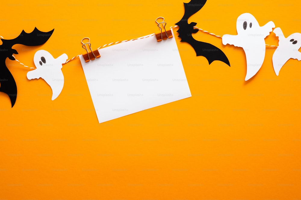 Happy halloween concept. Halloween decorations, blank paper card on clips, bats, ghosts on orange background. Halloween party greeting card mockup with copy space. Flat lay, top view, overhead.