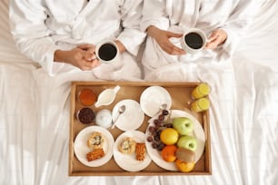 Top view of couple wearing white bath robes lying in the bed. Their breakfast is on the tray in the bed. Travelling together concept. Horizontal shot
