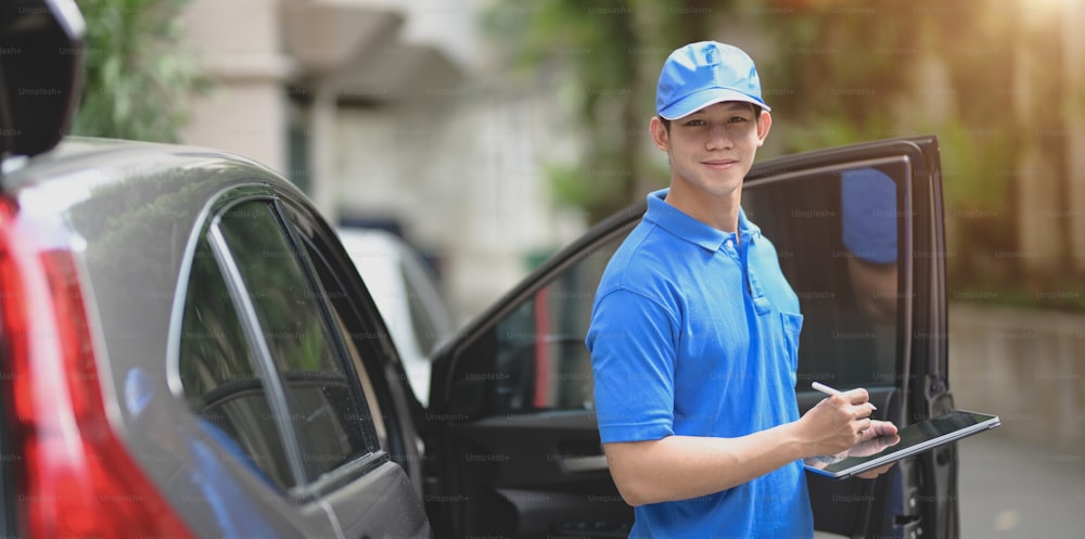 Delivery man checking orders for customer and smiling to the camera while standing in front of his car