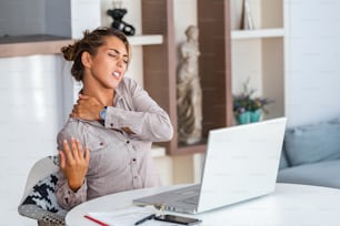 Portrait of young stressed woman sitting at home office desk in front of laptop, touching aching back with pained expression, suffering from backache after working on pc