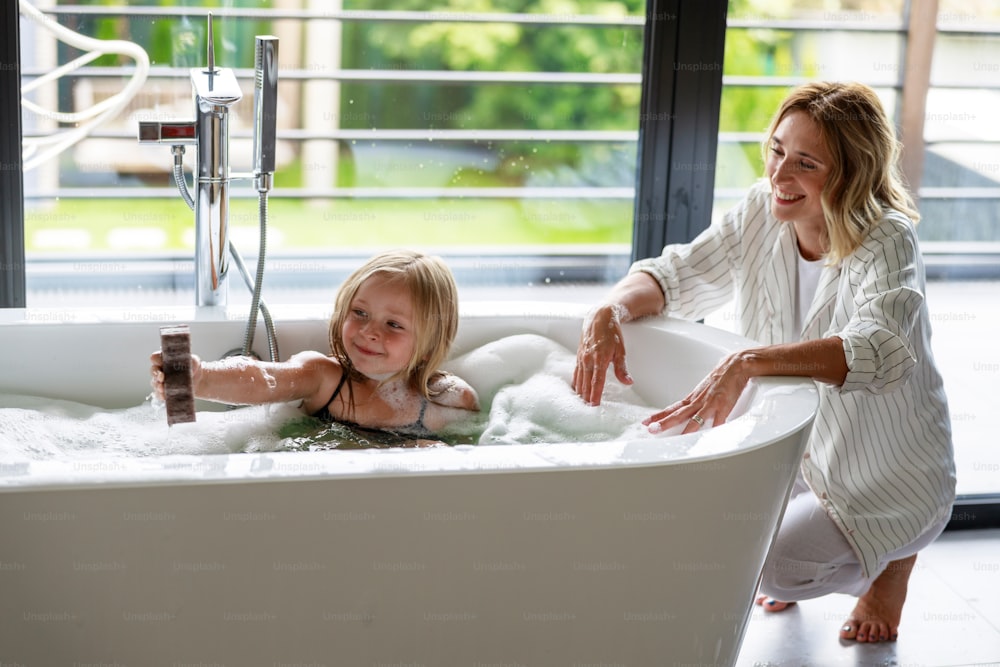 Smiling mom sitting near bath and looking after daughter stock photo
