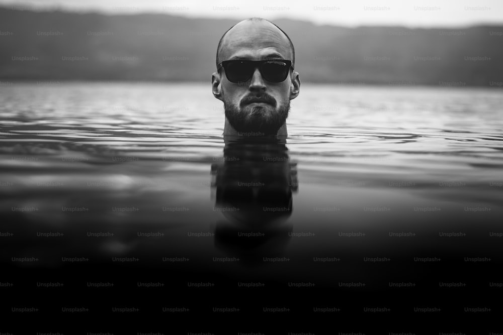 Brutal bearded man in sunglasses emerge in lake waves. Man head above water in lake in rainy foggy day, atmospheric moment. Wanderlust. Creative black and white photo