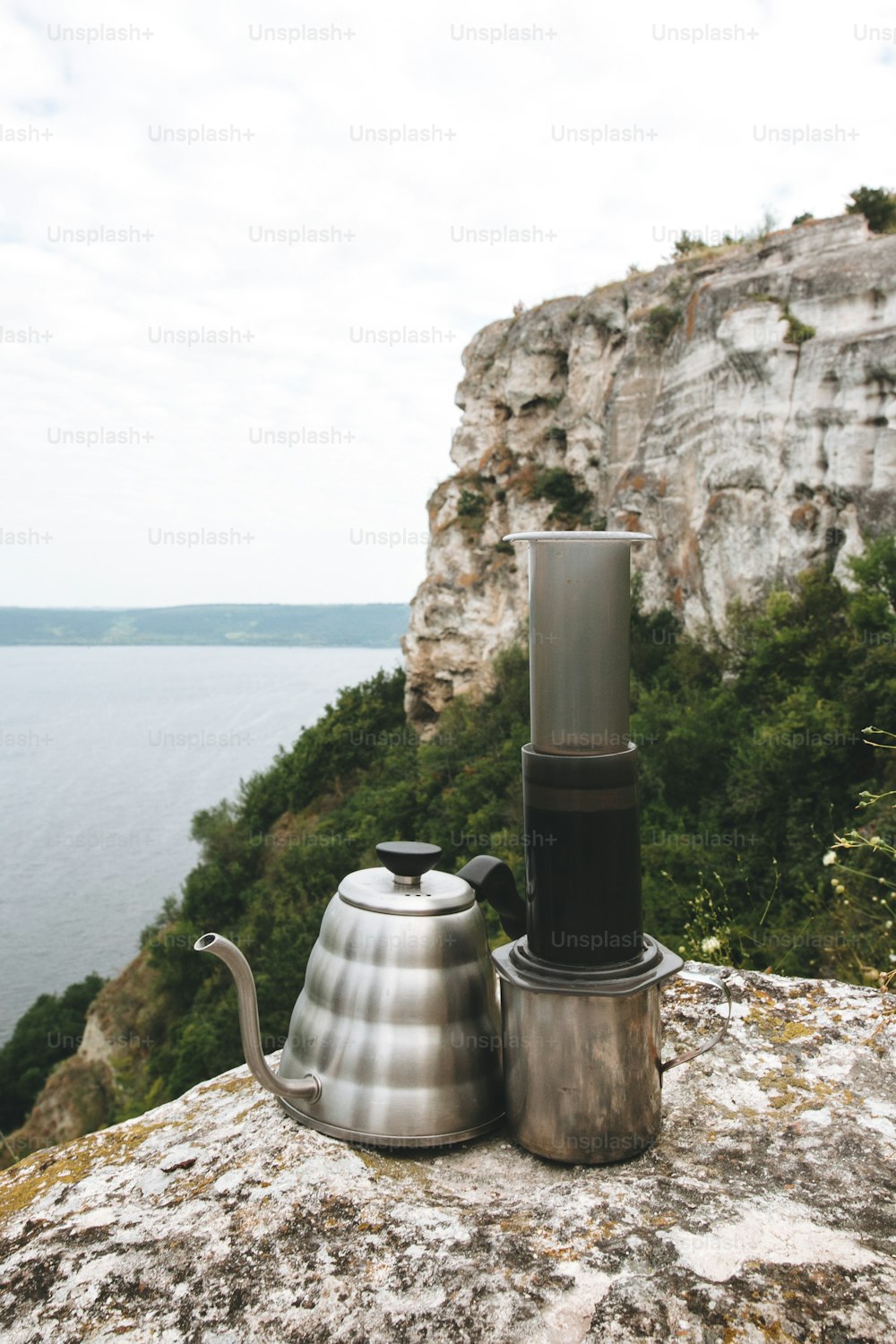 Steel kettle and aeropress on metal mug on cliff at lake, brewing alternative coffee at camping. Making hot drink at picnic outdoors. Trekking and hiking in mountains