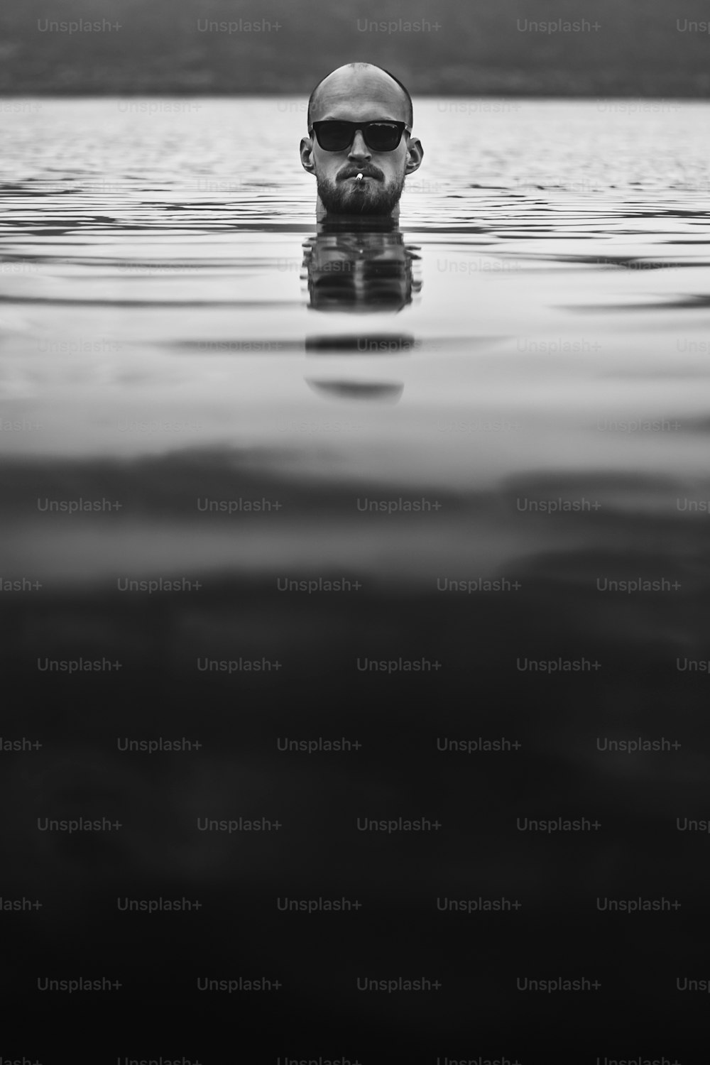 Brutale uomo barbuto con sigaretta e occhiali da sole emerge nel lago. Testa dell'uomo che fuma sopra l'acqua nel lago in una giornata nebbiosa piovosa, momento atmosferico. Voglia di viaggiare. Foto creativa in bianco e nero