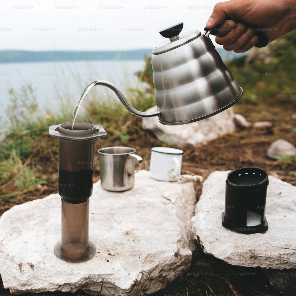 Brewing alternative coffee at camping. Traveler pouring hot water from steel kettle in aeropress on cliff at lake. Making hot drink at picnic outdoors. Trekking and hiking in mountains