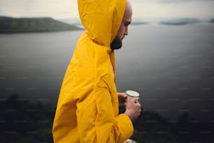 Wanderlust. Traveler in yellow raincoat holding metal mug and standing on cliff  in rainy windy day with view on lake. Hipster man hiking in Norway. Atmospheric moment