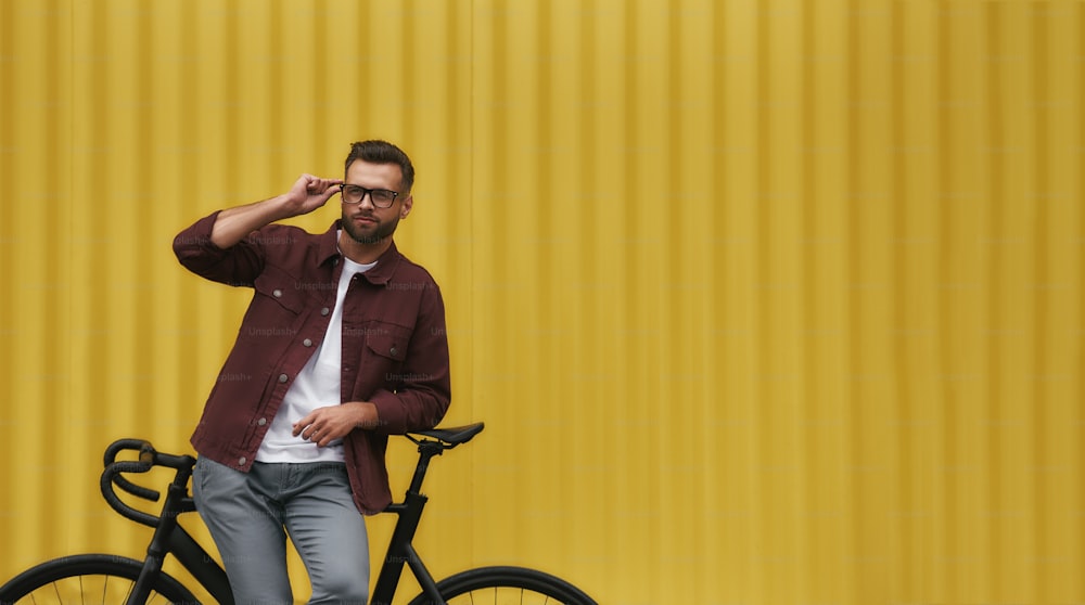 It was great ride! Handsome and young man with stubble in casual clothes adjusting his eyeglasses and leaning at his bicycle while standing against yellow wall outdoors. Weekend activity. City life