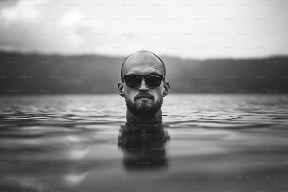 Un brutal hombre barbudo con gafas de sol emerge en las olas del lago. Cabeza del hombre por encima del agua en el lago en el día lluvioso y brumoso, momento atmosférico. Wanderlust. Foto creativa en blanco y negro