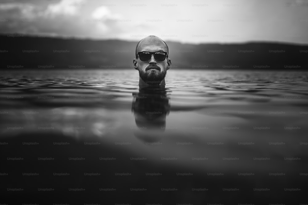 Brutal bärtiger Mann mit Sonnenbrille taucht in Seewellen auf. Mann Kopf über Wasser im See in regnerischen nebligen Tag, atmosphärischer Moment. Fernweh. Kreatives Schwarz-Weiß-Foto