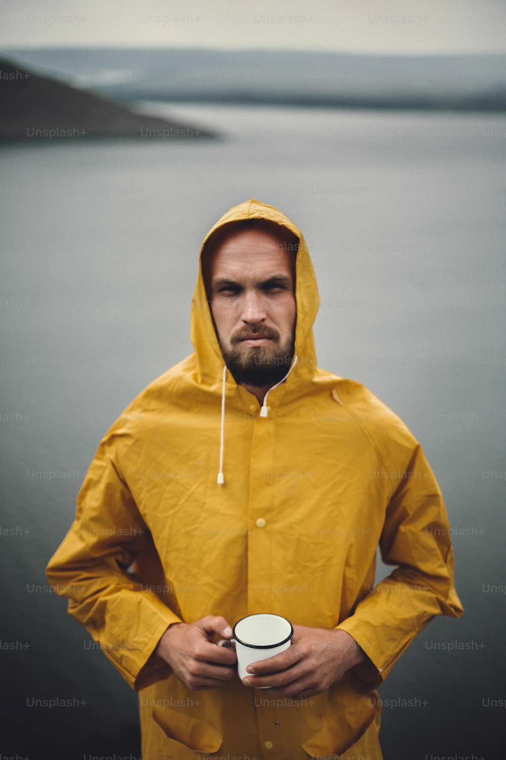 Brutal bearded man in yellow raincoat holding metal mug and traveling on cliff in rainy windy day with view on lake. Atmospheric moment. Wanderlust. Hipster man hiking in Norway