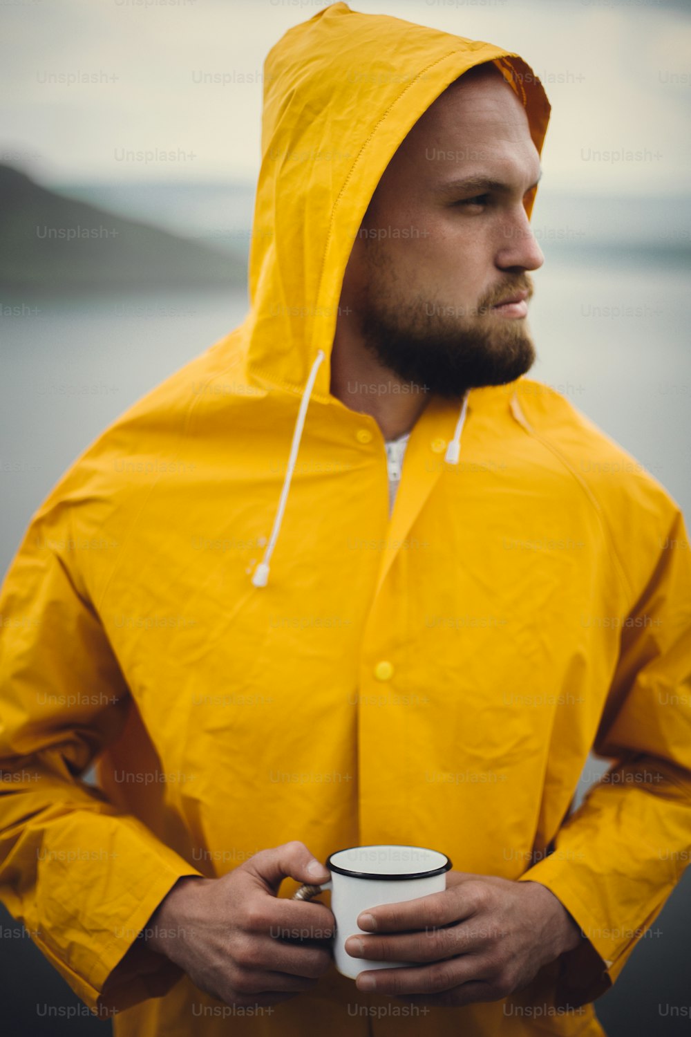 Traveler in yellow raincoat holding metal mug and standing on cliff  in rainy windy day with view on lake. Wanderlust and travel. Hipster man hiking in Norway. Atmospheric moment