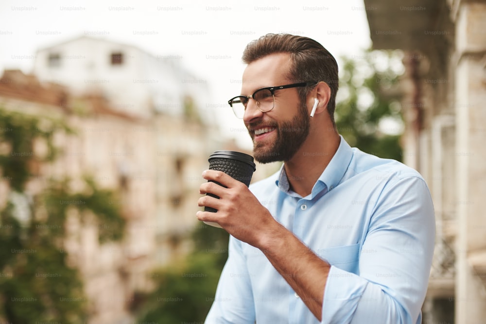 Young and handsome bearded man in eyeglasses and headphones holding cup of coffee and talking with friend while standing at the office balcony