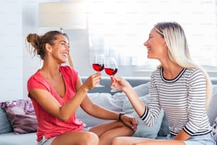 Two attractive girls, cheerful best friends having fun and drinking red wine at home. Two glasses of white wine in hands