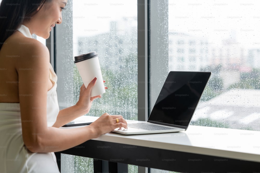 Young business woman using laptop computer while sitting at cafe table next to office windows.