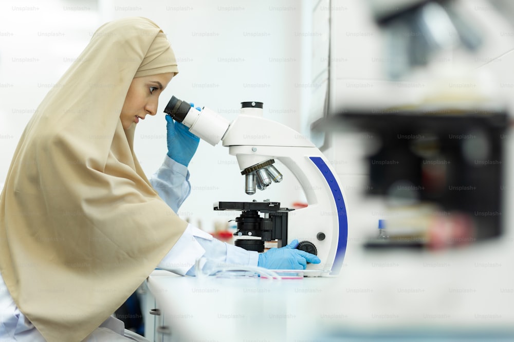 気にしないで。真面目なイスラム教徒の科学者が半姿勢で座り、顕微鏡で作業している