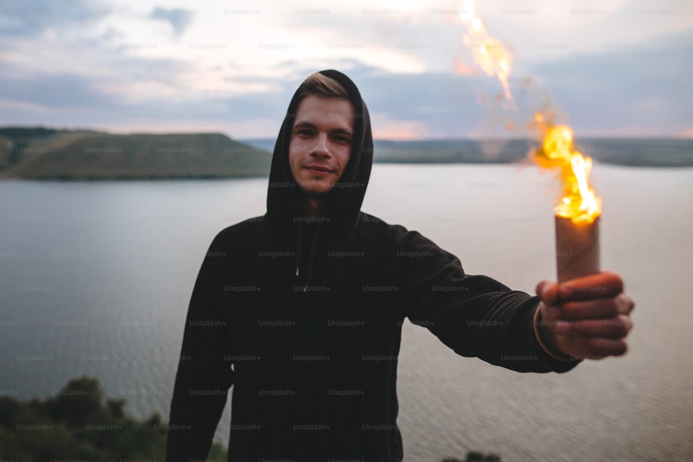 Ultras hooligan holding flare torch with burning fire in hand while standing on top of rock mountain with amazing sunset view on river . Atmospheric moment. Copy space