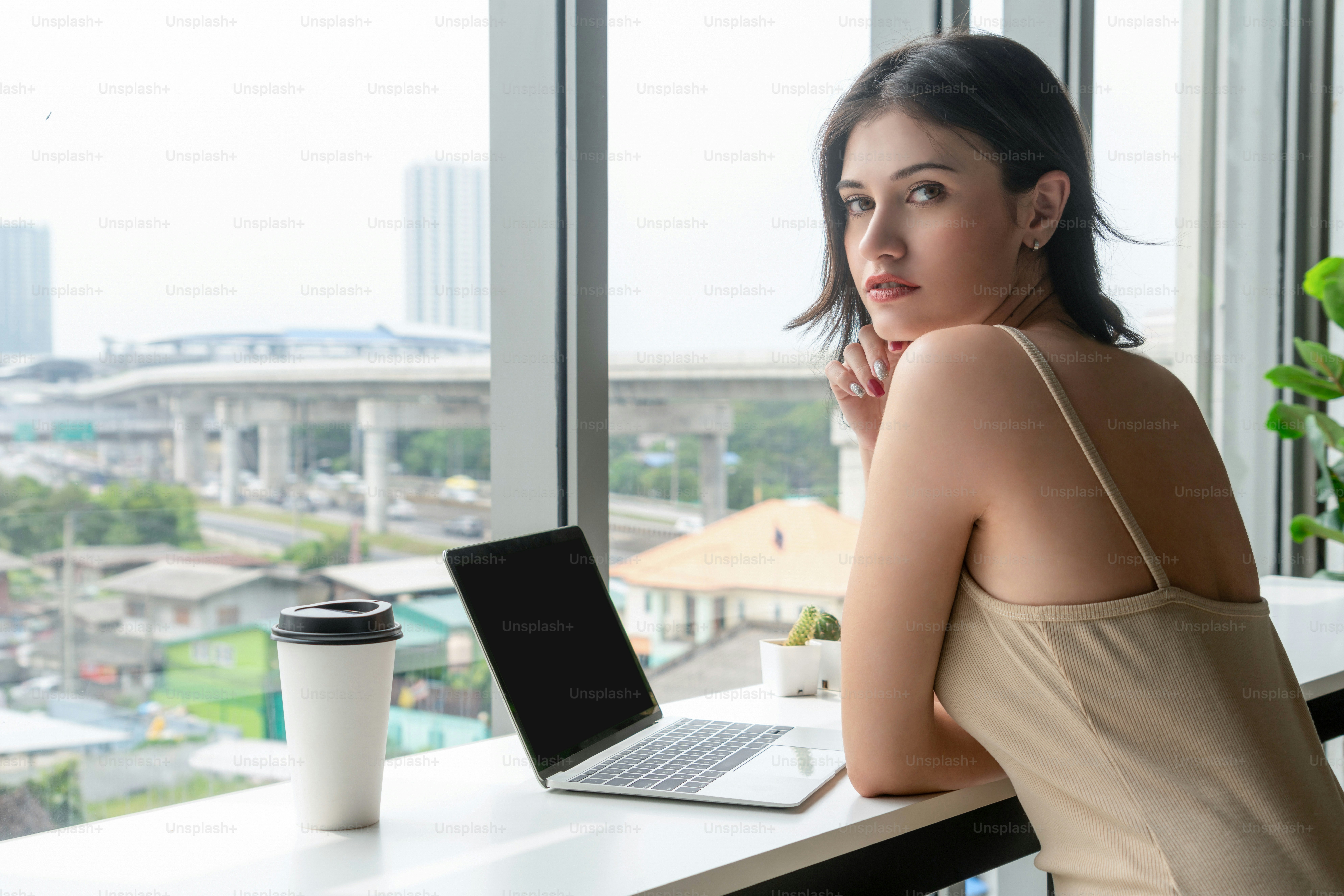 Junge Geschäftsfrau, die einen Laptop benutzt, während sie am Cafétisch neben den Bürofenstern sitzt.