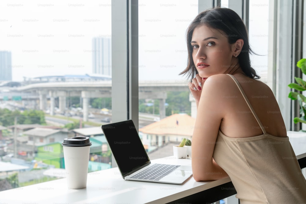 Young business woman using laptop computer while sitting at cafe table next to office windows.