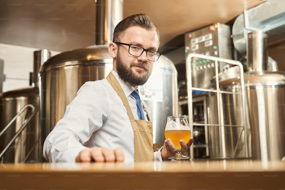 Young bearded brewer looking at camera and smiling while keeping glass of golden ale in hand. Man wearing white shirt and apron standing in brewery and examining beer. Concept of production.