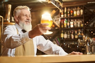 Enfoque selectivo de hombre alegre con camisa blanca y delantal de pie sobre la barra y sosteniendo un vaso de cerveza ligera. Hombre mirando hacia adelante y sonriendo mientras brinda con una taza. Concepto de cervecería.