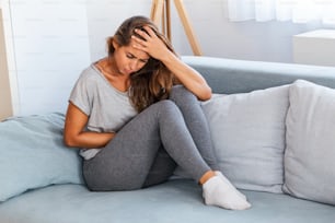 View of young woman suffering from stomachache on sofa at home. Woman sitting on bed and having stomach ache. Young woman suffering from abdominal pain while sitting on sofa at home