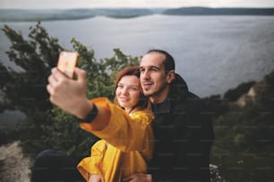 Happy hipster couple taking selfie on phone on top of rock mountain with beautiful view on river. Tourist couple hugging on windy cliff and making photo. Travel and wanderlust. Copy space