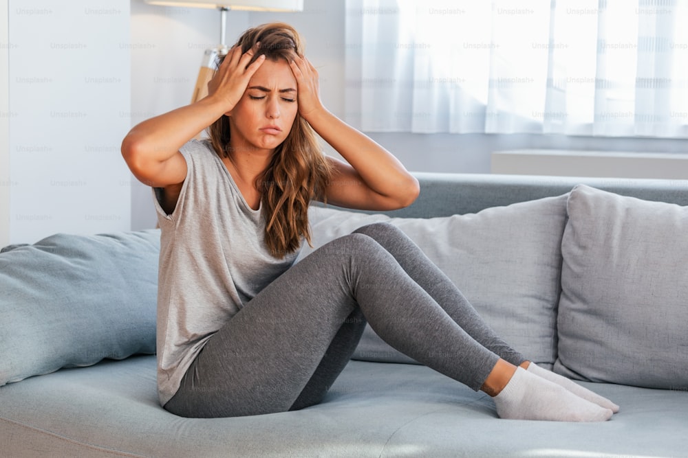 Portrait of an attractive woman sitting on a sofa at home with a headache, feeling pain and with an expression of being unwell.