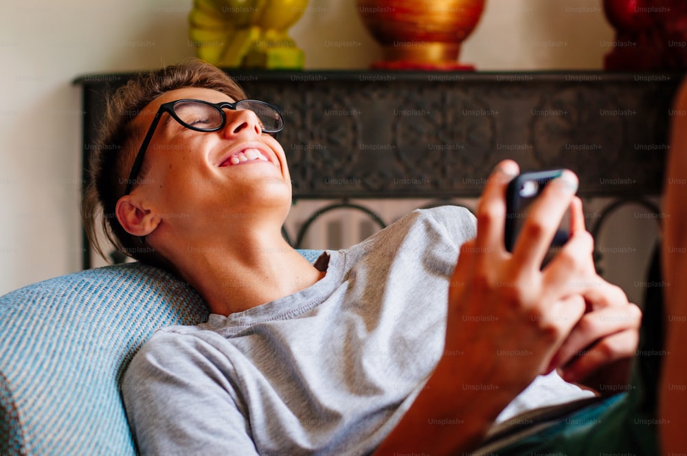 Concetto di Generazione Z. Ragazzo adolescente che ride usando lo smartphone che si gode la comunicazione seduto sulla sedia nel soggiorno.