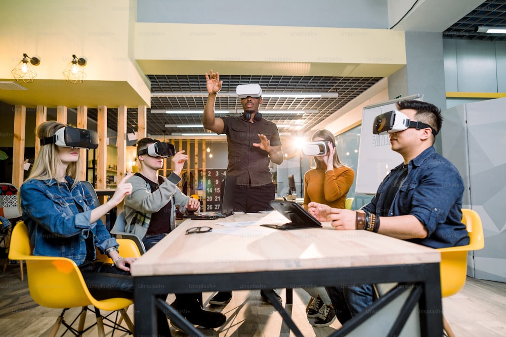 Testando jogos. Jovem africano usando fone de ouvido de realidade virtual e gesticulando enquanto estava de pé na mesa no escritório criativo junto com quatro amigos multiétnicos.