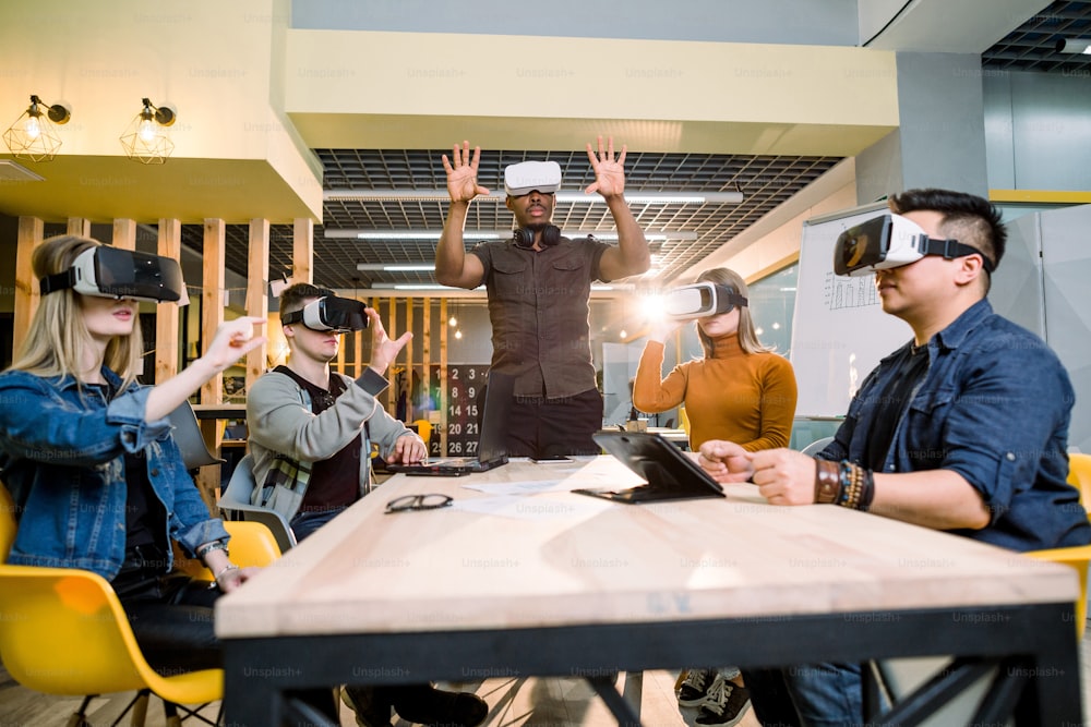 Un grupo de amigos y su hombre africano se prueban las gafas de realidad virtual mientras están sentados en la mesa de la oficina. Realidad virtual con personas que se divierten juntas con gafas de auriculares portátiles.