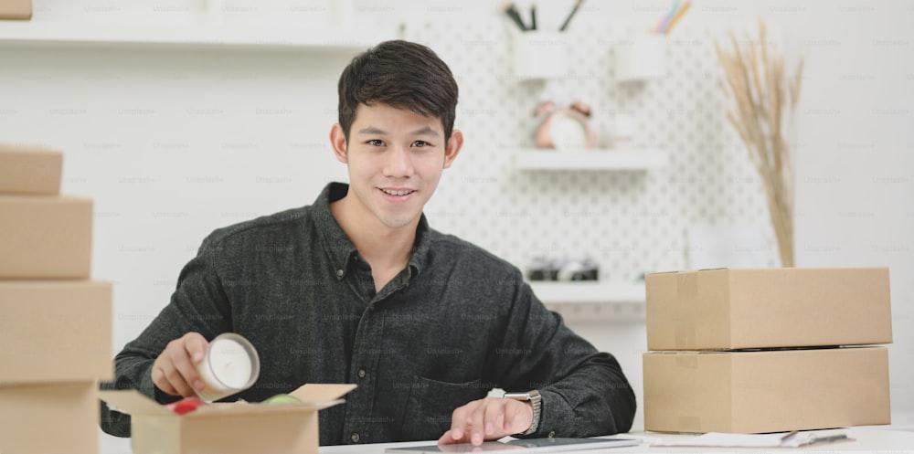 Jeune homme propriétaire d’une petite entreprise préparant et emballant les produits pour les clients à la maison et souriant à la caméra