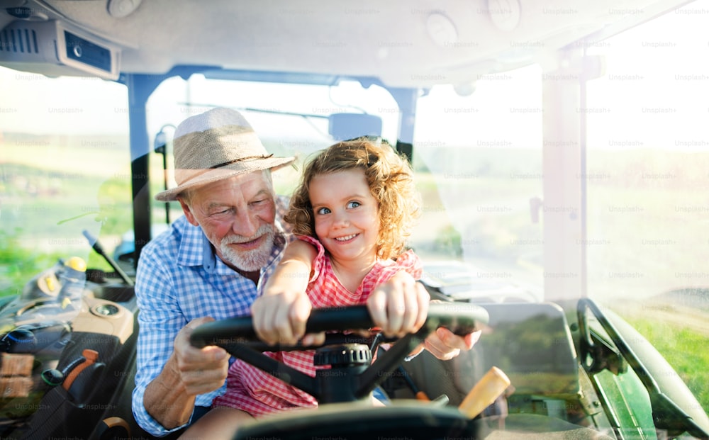 Senior Landwirt mit kleiner Enkelin, die im Traktor sitzt und fährt. Durch Glas geschossen.